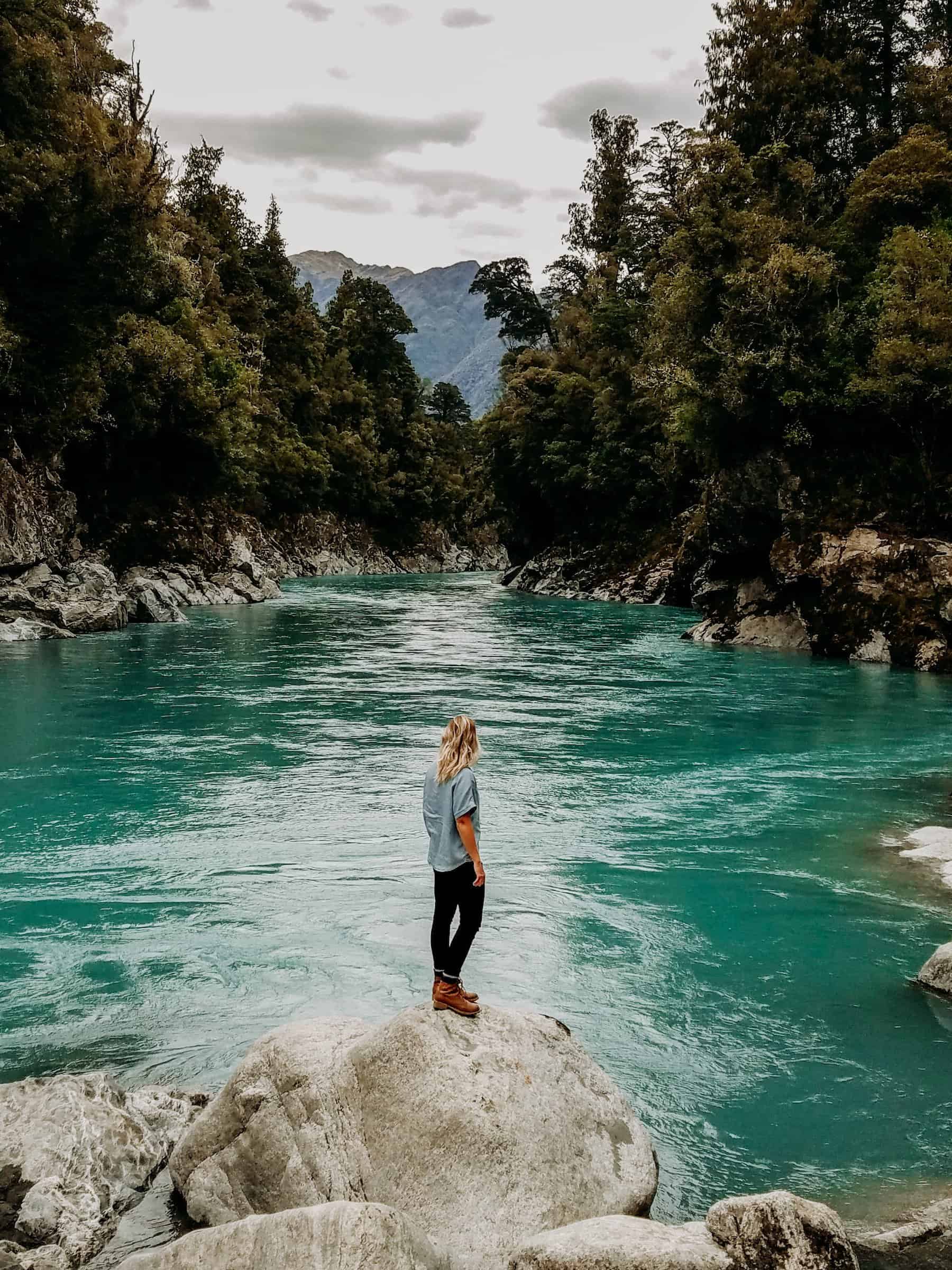A Hiker Standing On a Rocky Outcrop At The Hokitika Gorge Looking Out To The Turquoise Blue Glacial River Fringed By White Granie Rock And Native Podocard Rainforest On The West Coast Of New Zealand