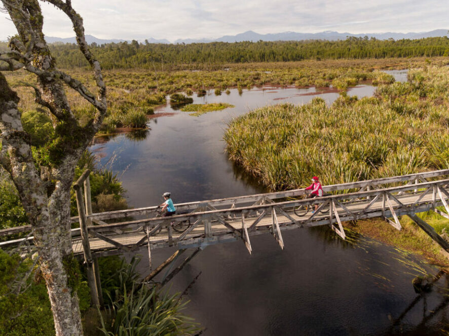 Mountain Bikers Crossing A Suspension Bridge Over The Lake Mahinapua Lagoon On The Mananui Tramway